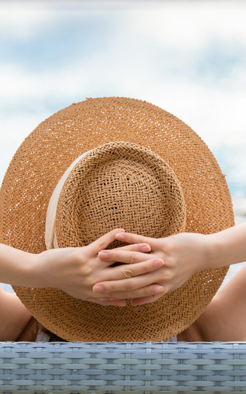 View of woman in sun hat with hands behind her head relaxing at swimming pool